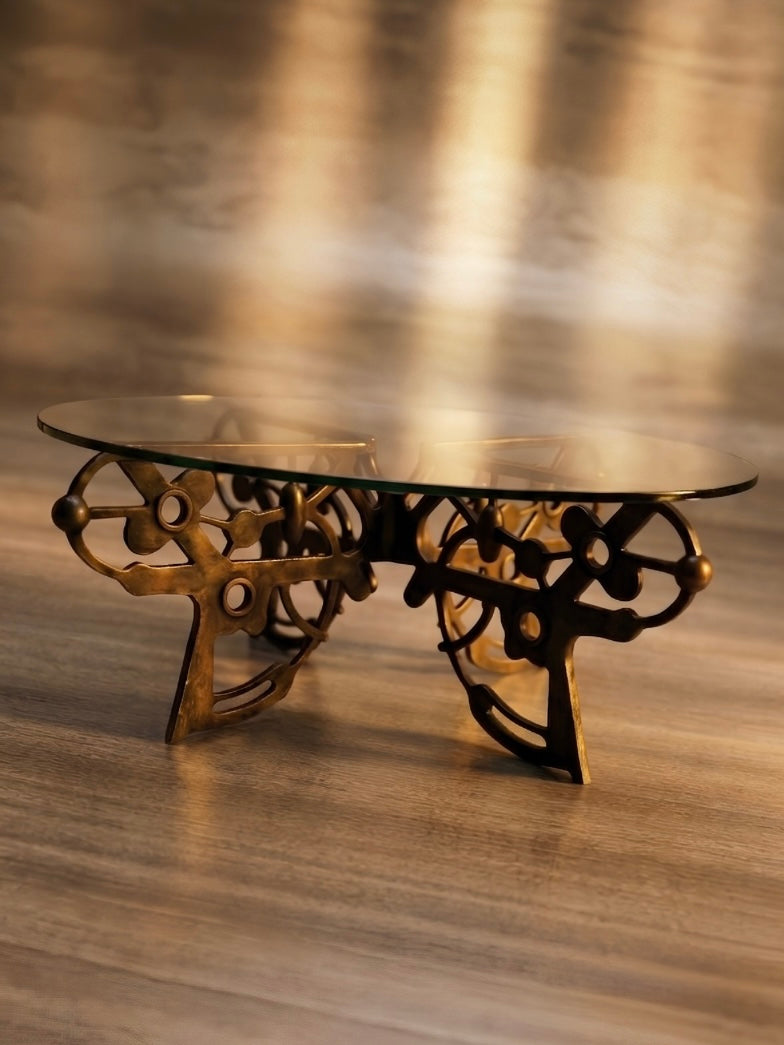 An ornate coffee table with a clear glass top and an intricate bronze-finished metal base, set on a wooden floor against a softly blurred background.