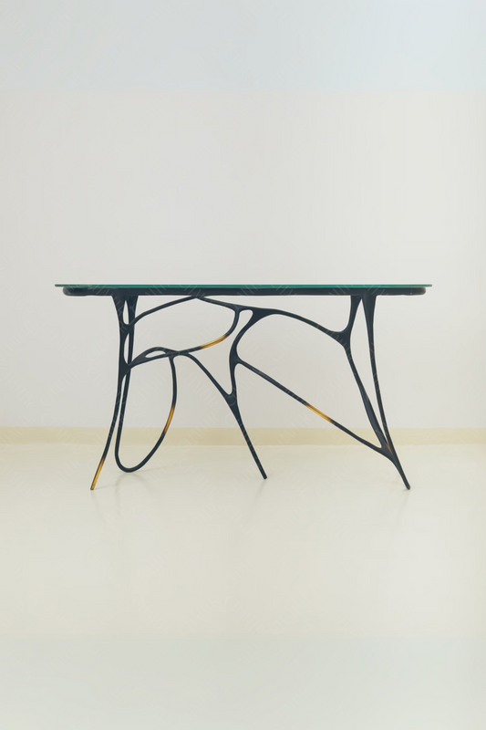 Black brass luxury handmade furniture table with organic design and glass top, perfect for modern luxury decor, on a light wooden floor with white wall background.