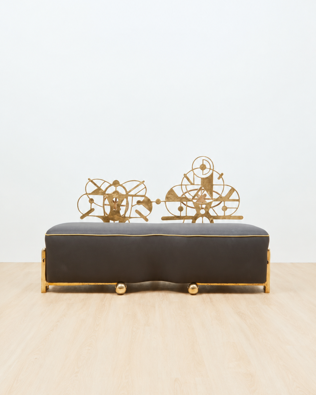 A sculptural couch with a dark charcoal gray upholstered seat and gold metal frame, featuring decorative gold wire sculptures on top, set against a plain white wall and light wood floor.