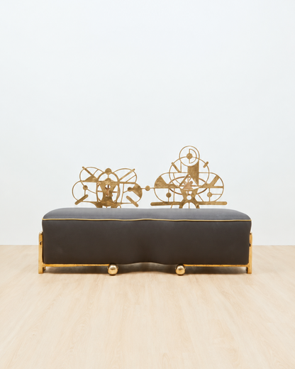 A sculptural couch with a dark charcoal gray upholstered seat and gold metal frame, featuring decorative gold wire sculptures on top, set against a plain white wall and light wood floor.