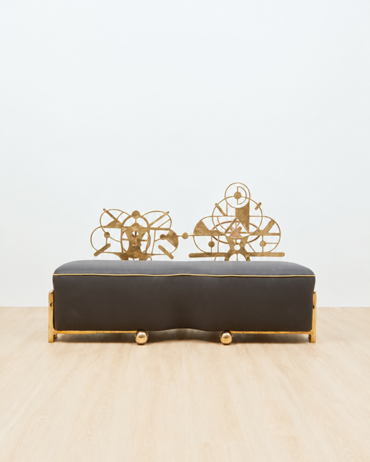 A sculptural couch with a dark charcoal gray upholstered seat and gold metal frame, featuring decorative gold wire sculptures on top, set against a plain white wall and light wood floor.