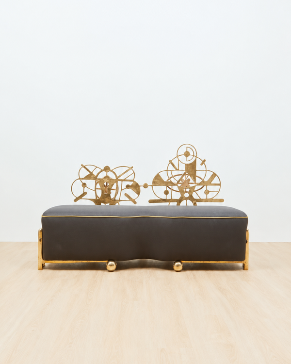 A sculptural couch with a dark charcoal gray upholstered seat and gold metal frame, featuring decorative gold wire sculptures on top, set against a plain white wall and light wood floor.
