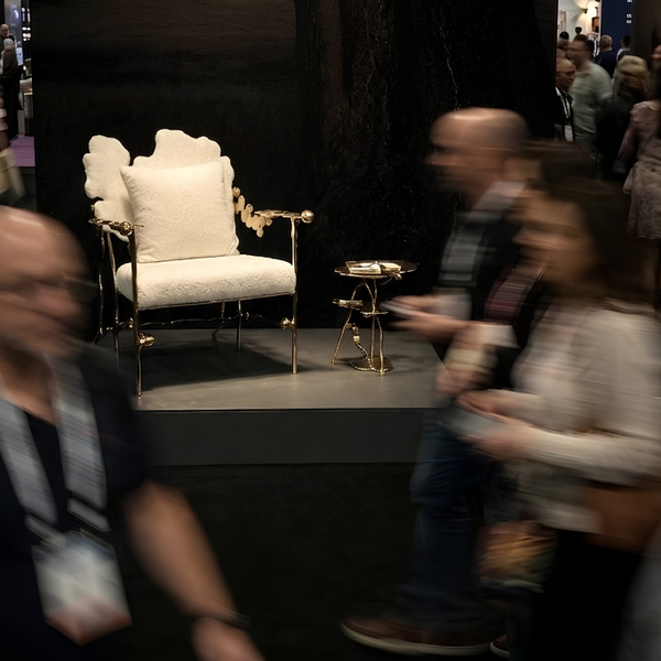 A white upholstered armchair with gold metal arms and legs sits on a raised platform at a trade show, with blurred people walking in the foreground and background.