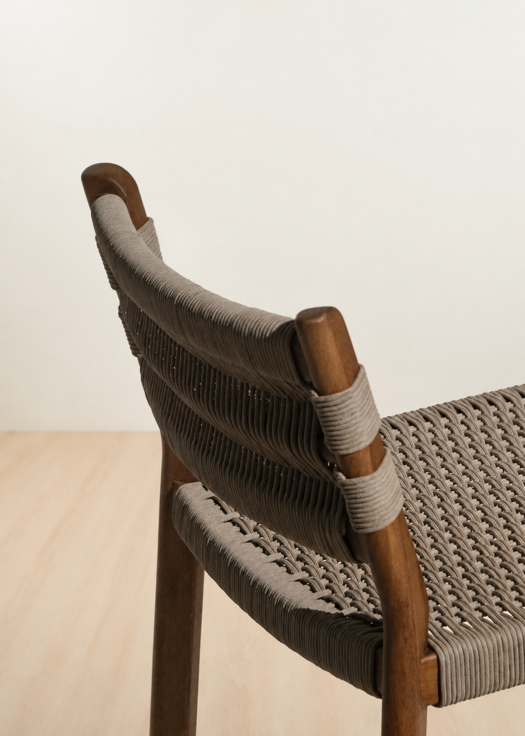Close-up of a modern wooden chair with woven gray rope seating and backrest, shown in a minimalist indoor setting with a light wall and pale wood floor.