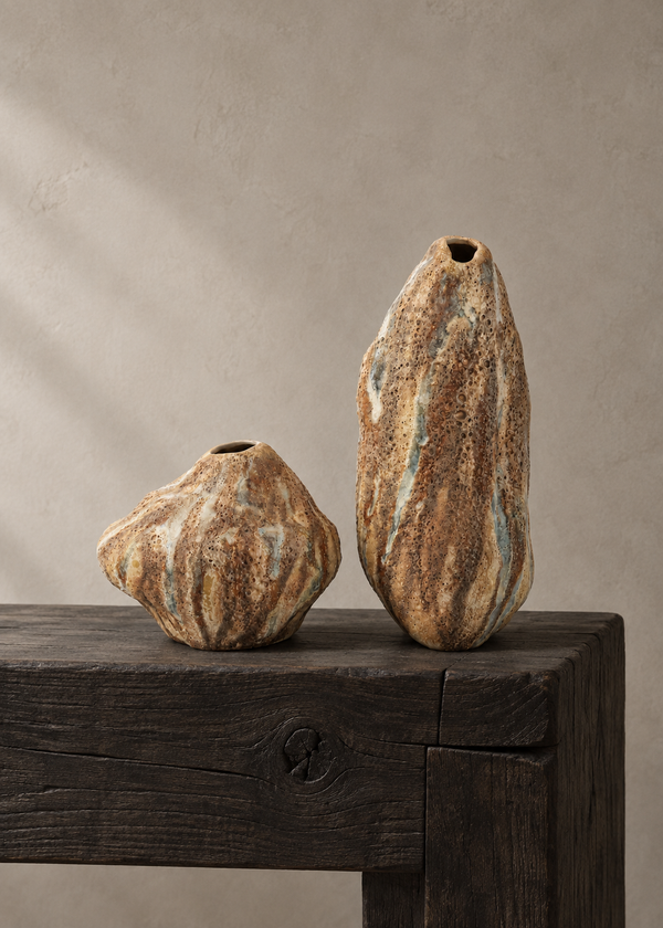 Two organic-shaped ceramic vases with textured, earthy brown and beige glazes, one short and bulbous and one tall and narrow, displayed on a dark rustic wooden table against a neutral beige wall.