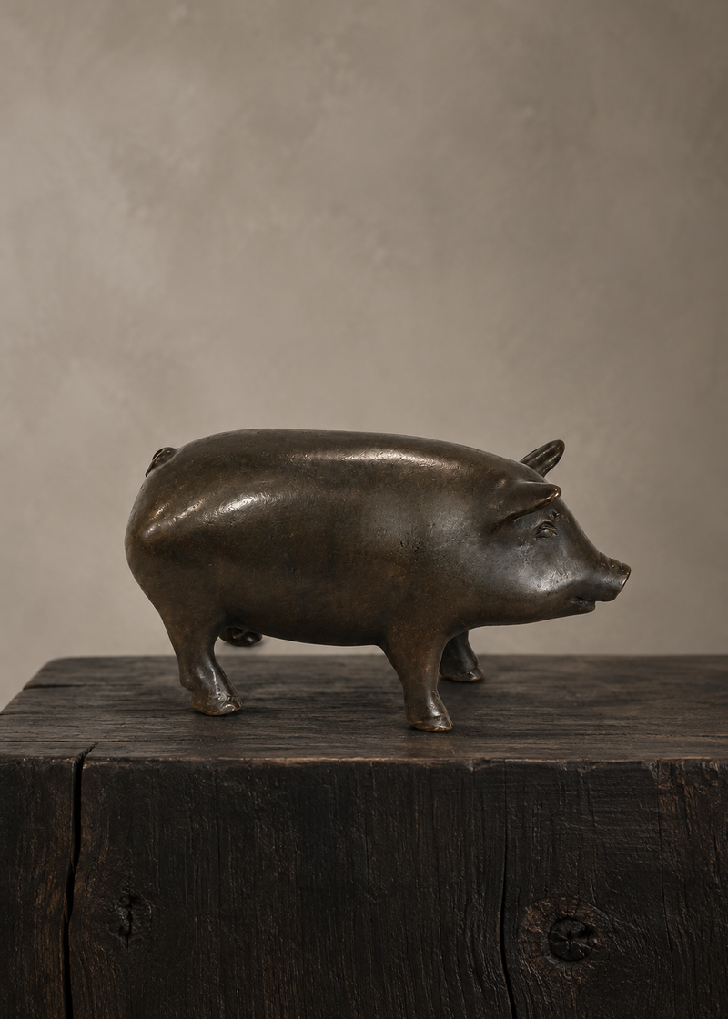 A dark bronze pig figurine standing on a rustic dark wooden block against a textured warm gray studio background.