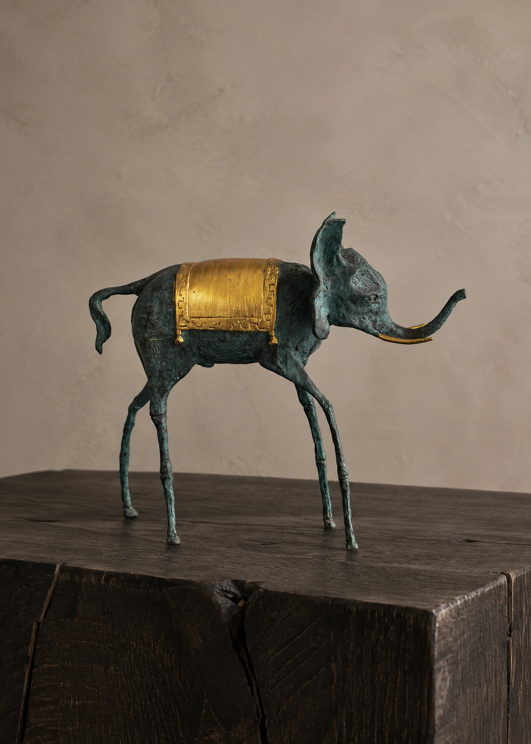 A small elephant bronze figurine with a dark green patina finish and a shiny gold saddle, standing on a rustic dark wooden block against a textured beige wall.