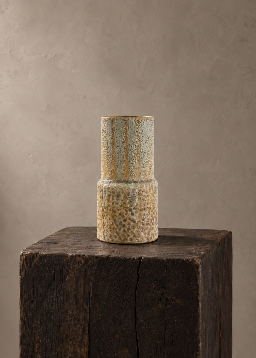 A two-tiered ceramic vase with a textured, speckled beige and brown glaze, displayed on a rustic dark wood block against a neutral textured beige background.