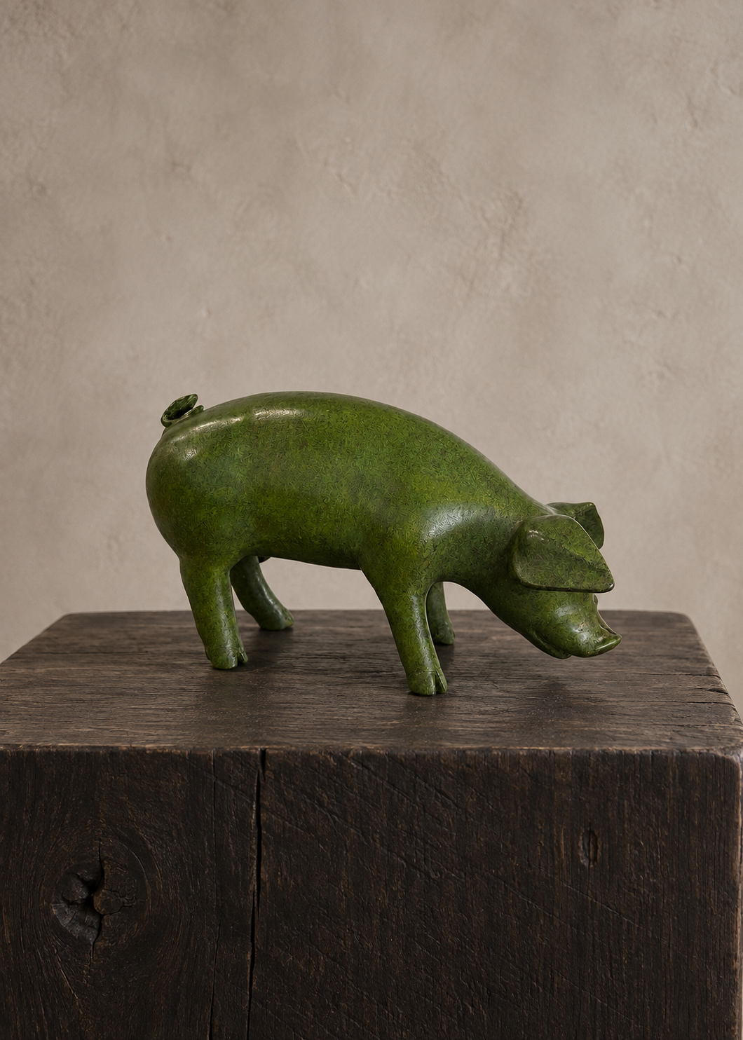 A green pig-shaped bronze sculpture or container with a glossy finish, standing on a dark wooden block against a textured beige wall.