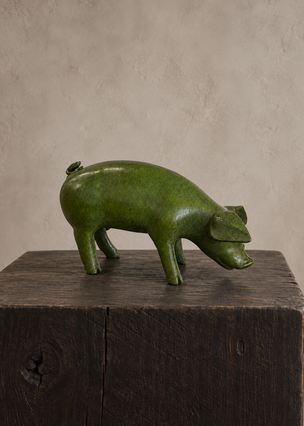 A green pig-shaped bronze sculpture or container with a glossy finish, standing on a dark wooden block against a textured beige wall.