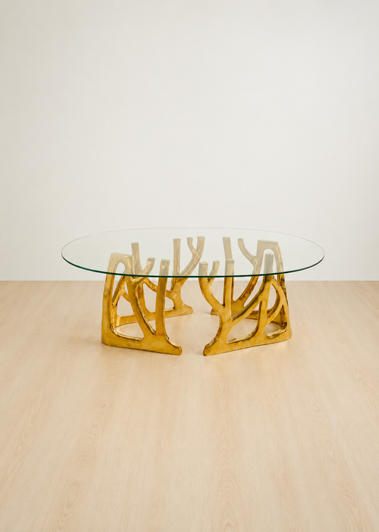 An oval glass-top coffee table with a sculptural, branching polished brass base, set on a light wood floor against a plain off-white wall.