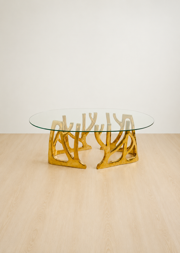 An oval glass-top coffee table with a sculptural, branching polished brass base, set on a light wood floor against a plain off-white wall.