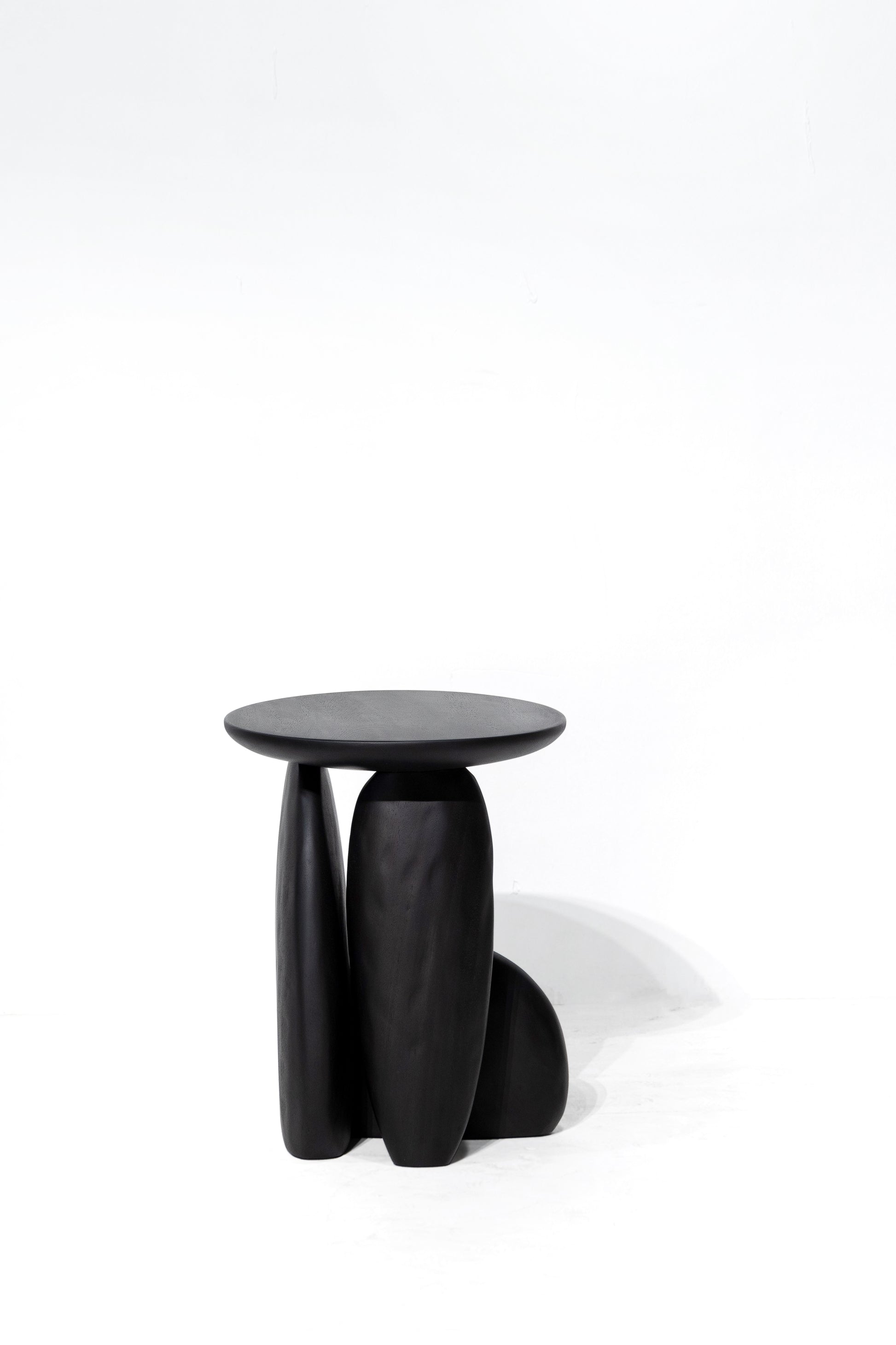 Pebble stool. A minimalist black sculptural stool with a round top and three organic-shaped legs, photographed on a seamless white studio background.