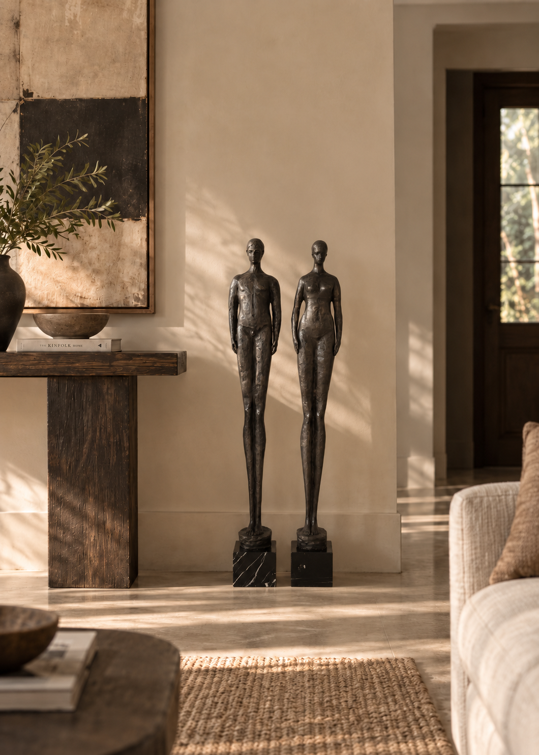 Two dark bronze human figure sculptures standing on black marble bases in a sunlit, modern living room interior.