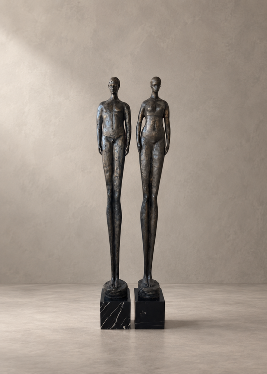 Two tall abstract female nude statues in dark bronze finish standing on black marble bases, photographed against a neutral textured beige wall and floor.