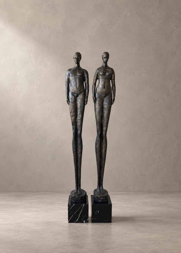 Two tall abstract female nude statues in dark bronze finish standing on black marble bases, photographed against a neutral textured beige wall and floor.