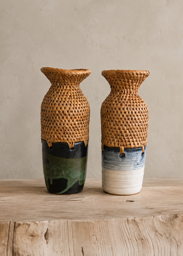 Two handmade woven rattan vases with glazed ceramic bases, displayed on a rustic wooden table against a textured beige plaster wall.