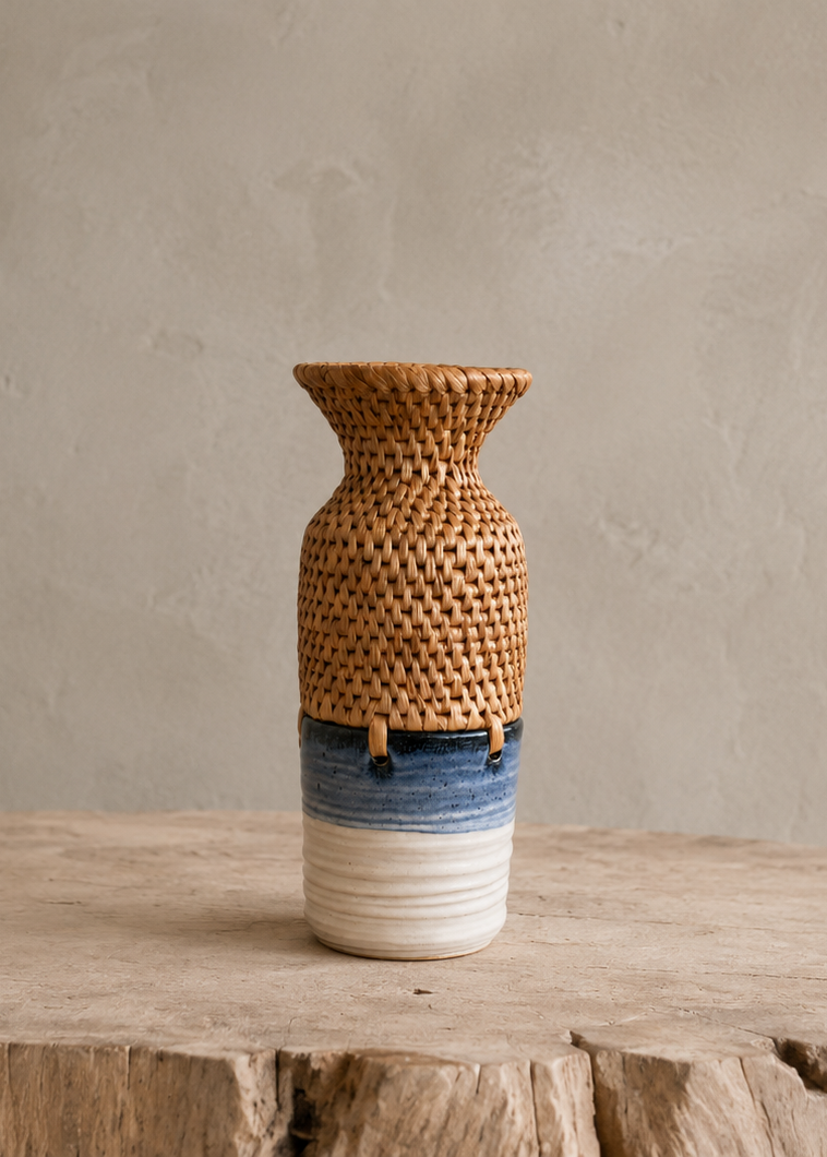 A small decorative vase with a woven natural rattan upper section, a blue band, and a white ribbed base, displayed on a rustic wood slice against a textured beige wall.