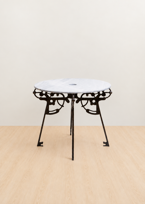 A round outdoor bar table featuring a white marble top and an ornate black metal frame with four legs, set against a minimalist beige wall and light wood floor.