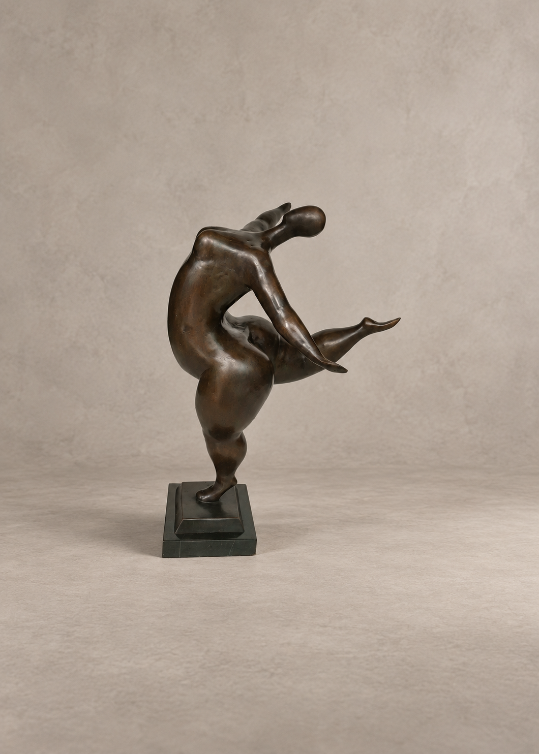 A bronze sculpture of a dancing lady in an abstract, stylized pose, mounted on a dark rectangular base against a neutral textured beige background.
