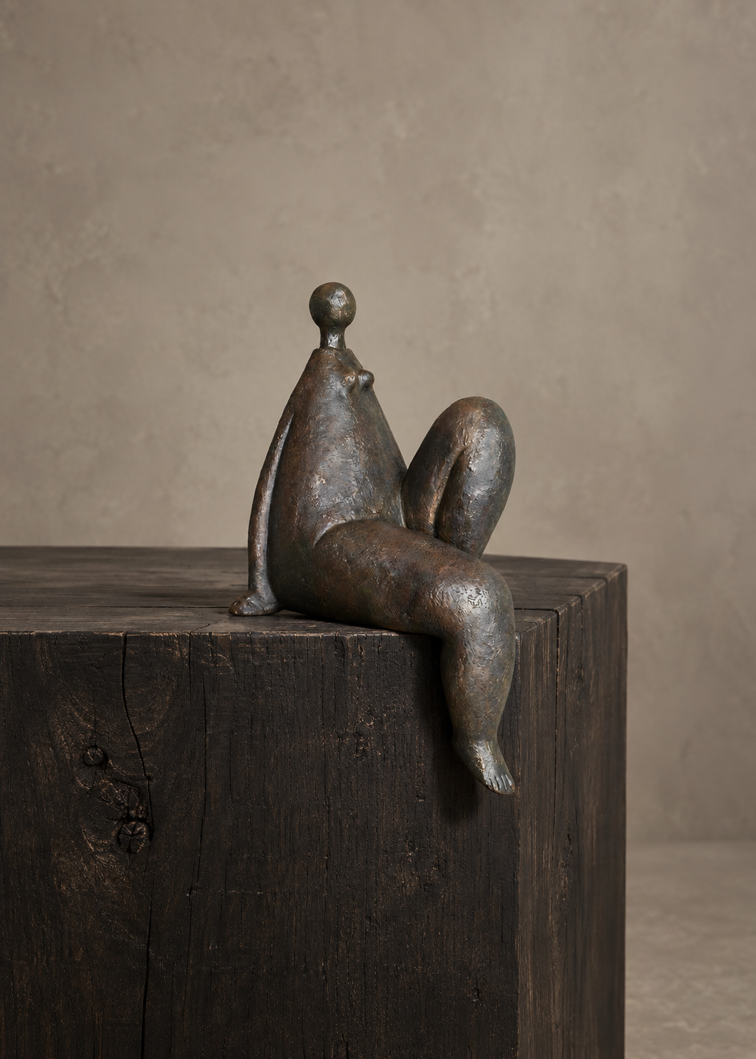 A small abstract bronze sculpture of a seated human figure resting on the corner of a dark wooden block against a warm beige textured wall.