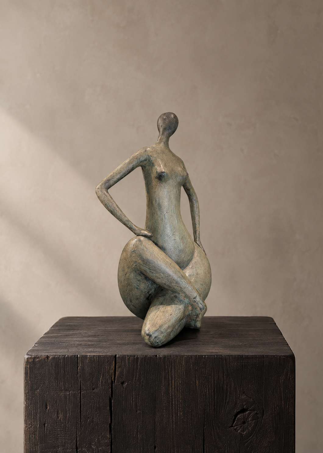 A bronze sculptural figurine of a seated nude female form in muted gray-green tones, displayed on a dark wooden pedestal against a warm beige textured wall.