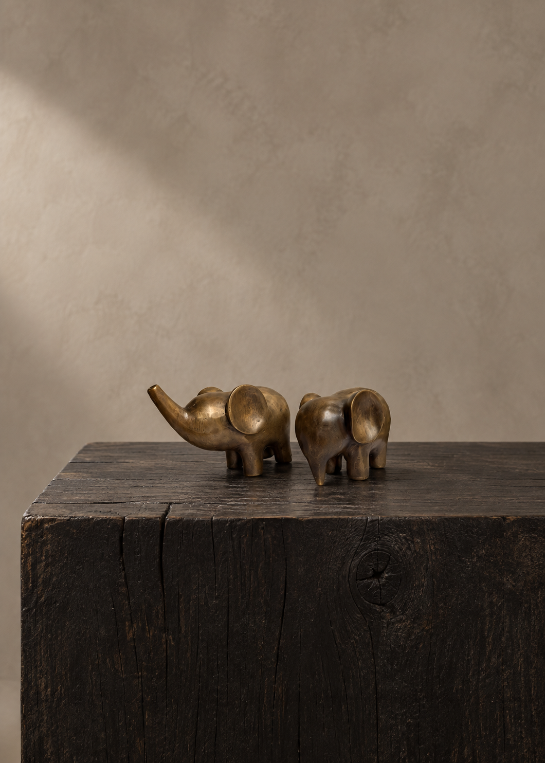 Two small bronze elephant figurines in a warm gold finish, displayed on top of a dark rustic wooden block against a textured beige wall.